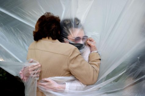 Two people embrace in the only way two people can during a pandemic: through a plastic sheet and wearing face masks.